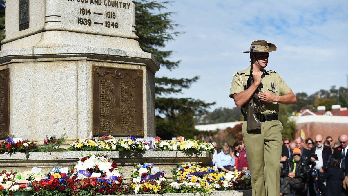 A solider holding a rifle standing in front of a war memorial which has many colourful wreaths at its base. A crowd of people are looking on in the background.