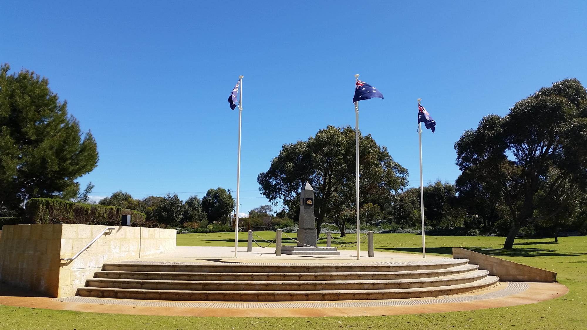  war memorial located in an open park with large trees in the background and three flagpoles with Australian flags flying.